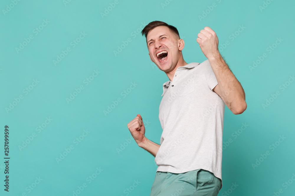 Portrait of overjoyed screaming young man in casual clothes clenching fists like winner isolated on blue turquoise background in studio. People sincere emotions, lifestyle concept. Mock up copy space. Portrait of overjoyed screaming young man in casual clothes clenching fists like winner isolated on blue turquoise background in studio. People sincere emotions, lifestyle concept. Mock up copy space.