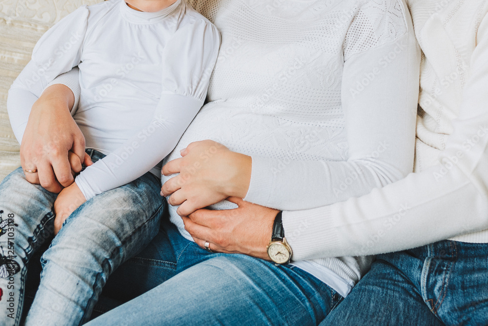 Pregnant woman sitting with her husband and an older child. The family are wearing jeans and white sweaters. Concept of pregnancy and expecting a baby. Pregnant woman sitting with her husband and an older child. The family are wearing jeans and white sweaters. Concept of pregnancy and expecting a baby.