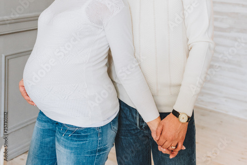 Pregnant woman holding hands with her husband. The couple are wearing jeans and white sweaters. Concept of pregnancy and expecting a baby.