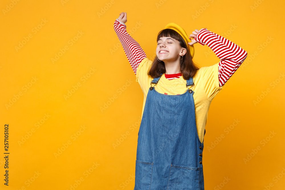 Joyful girl teenager in french beret, denim sundress keeping eyes closed, spreading and rising hands isolated on yellow wall background. People sincere emotions, lifestyle concept. Mock up copy space. Joyful girl teenager in french beret, denim sundress keeping eyes closed, spreading and rising hands isolated on yellow wall background. People sincere emotions, lifestyle concept. Mock up copy space.