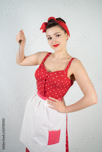 dangerous pin up girl housewife in red vintage polka dot dress stands with a huge kitchen knife in her hands and angrily rejoices on white solid studio background