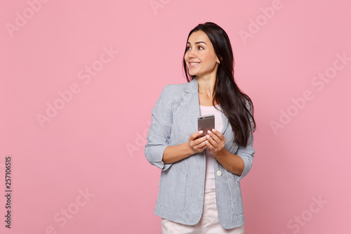 Smiling young woman in striped jacket looking aside using mobile phone, typing sms message isolated on pink pastel background in studio. People sincere emotions, lifestyle concept. Mock up copy space.