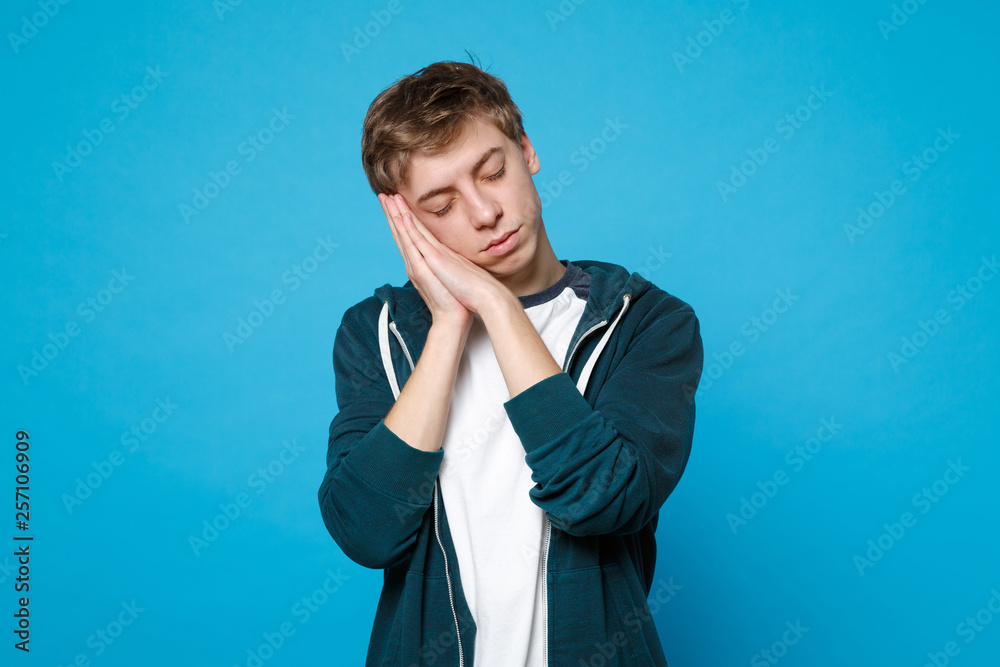 Portrait of relaxed young man in casual clothes keeping eyes closed, resting, sleaping with hands near face isolated on blue background. People sincere emotions lifestyle concept. Mock up copy space.