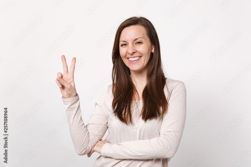 Portrait of smiling young woman in light clothes looking camera and showing victory sign isolated on white wall background in studio. People sincere emotions, lifestyle concept. Mock up copy space. - obrazy, fototapety, plakaty 