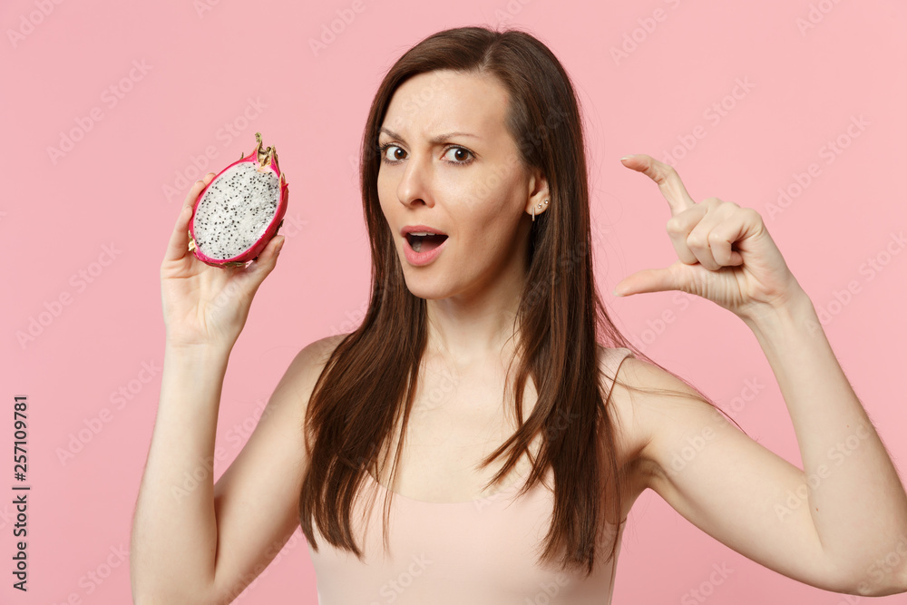 Puzzled young woman gesturing demonstrating size with workspace, holding fresh ripe pitahaya, dragon fruit isolated on pink pastel background. People vivid relax vacation concept. Mock up copy space.