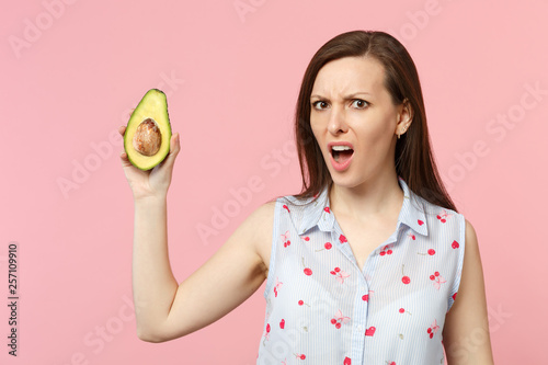 Concerned perplexed young woman in summer clothes holding half of fresh ripe green avocado fruit isolated on pink pastel background. People vivid lifestyle relax vacation concept. Mock up copy space.