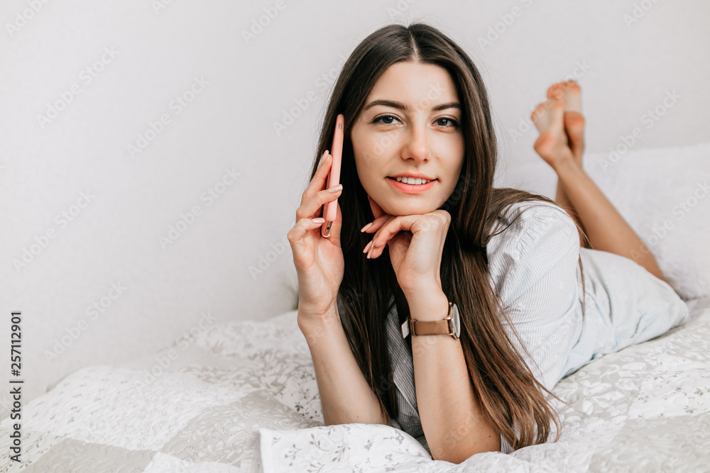 Young smiling woman lying in white bed and using a phone in her bedroom. Happy morning. Young smiling woman lying in white bed and using a phone in her bedroom. Happy morning.