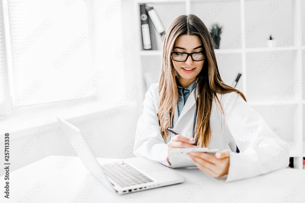 Smiling young female doctor working at the clinic , she is using a computer and writing medical reports. Smiling young female doctor working at the clinic , she is using a computer and writing medical reports.