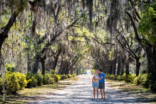 Street road landscape with oak trees path in Savannah, Georgia famous Bonaventure cemetery with Spanish moss and young happy romantic couple standing embracing love photo