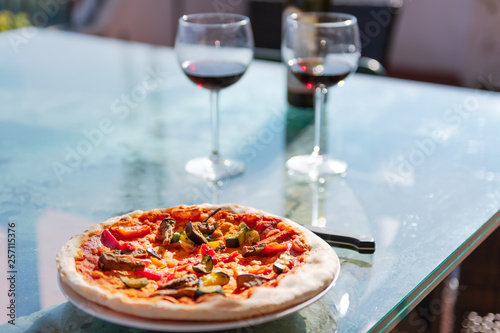 Fresh pizza on glass table terrace outside in Italy with tomato sauce vegetables and two glasses of red wine bottle
