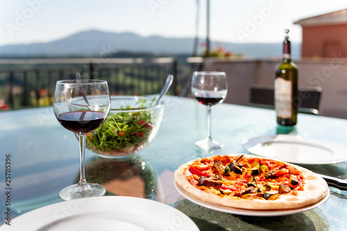 Fresh pizza and salad on glass table terrace outside in Italy with tomato sauce vegetables and two glasses of red wine bottle