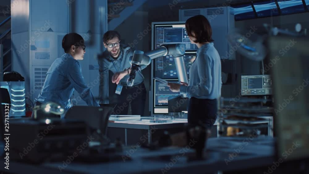 Diverse Team of Engineers with Laptop and a Tablet Analyse and Discuss How a Futuristic Robotic Arm Works and Moves a Metal Object. They are in a High Tech Research Laboratory with Modern Equipment. Diverse Team of Engineers with Laptop and a Tablet Analyse and Discuss How a Futuristic Robotic Arm Works and Moves a Metal Object. They are in a High Tech Research Laboratory with Modern Equipment.