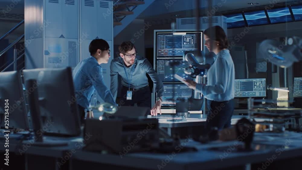 Diverse Team of Engineers with Laptop and a Tablet Analyse and Discuss How a Futuristic Robotic Arm Works and Moves a Metal Object. They are in a High Tech Research Laboratory with Modern Equipment.