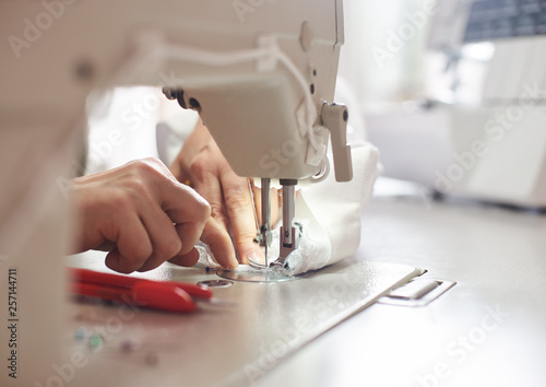 Woman hands stitching white fabric on professional manufacturing machine at workplace. Seamstress hands holding lace textile for dress production. Blurred background. Close up view of sewing process.