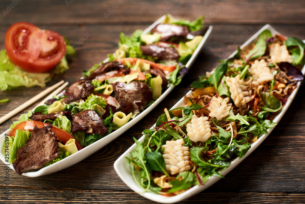 Hot Salads with squid, soybean sprouts, beef, avocado, rucola on recipe of Japanese cuisine in white plate. Chopsticks, tomato, lettuce, basil on wooden table serving in Asian tradition. Top view. Hot Salads with squid, soybean sprouts, beef, avocado, rucola on recipe of Japanese cuisine in white plate. Chopsticks, tomato, lettuce, basil on wooden table serving in Asian tradition. Top view.