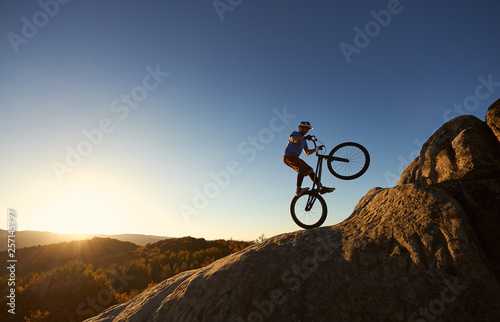 Silhouette of courageous cyclist balancing on back wheel on trial bicycle. Professional sportsman biker making acrobatic stunt on the top of mountain at sunset. Concept of extreme sport. Copy space