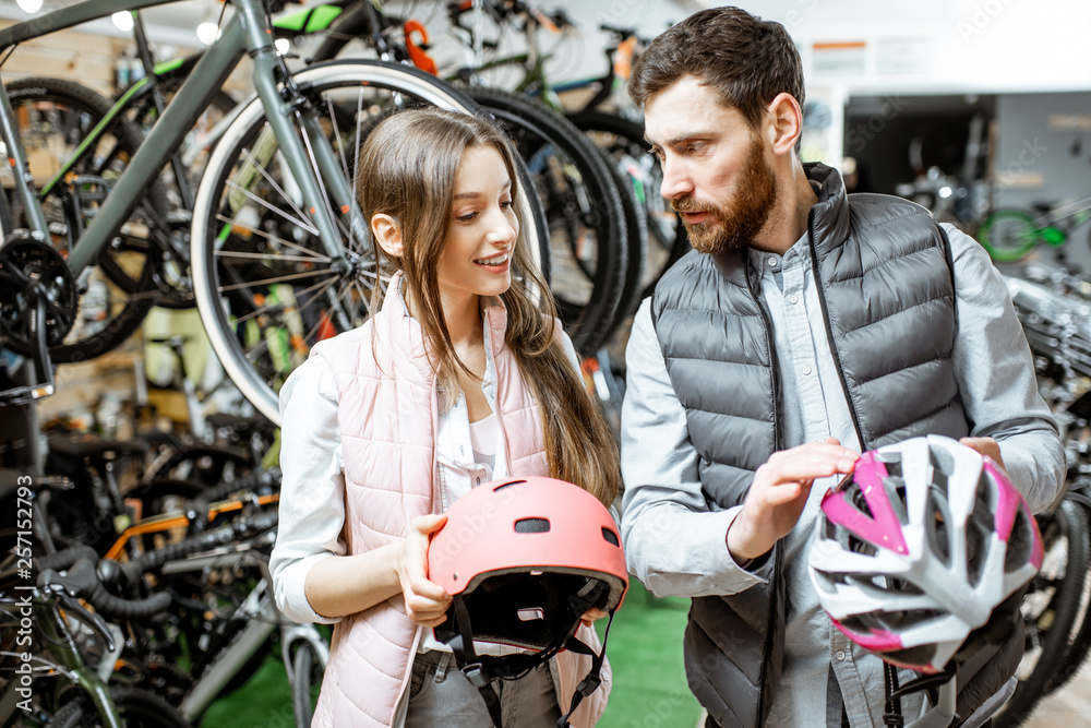 Young woman customer choosing protective helmet standing with salesman in the bicycle shop Young woman customer choosing protective helmet standing with salesman in the bicycle shop