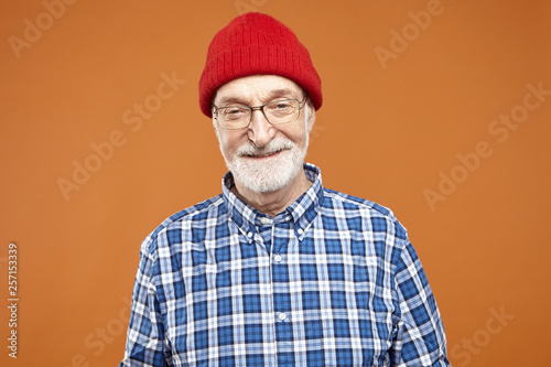 People, lifestyle, aging and positive human facial expressions. Portrait of cheerful handsome senior male pensioner posing at blank orange wall, wearing red hat and plaid shirt, smiling broadly