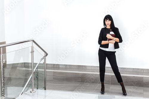 Young beautiful serious brunette woman with a note book dressed in a black business suit standing against the white wall in a office.