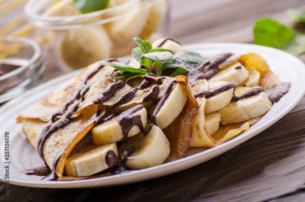 French crepes with chocolate sauce and banana in ceramic dish on wooden kitchen table French crepes with chocolate sauce and banana in ceramic dish on wooden kitchen table
