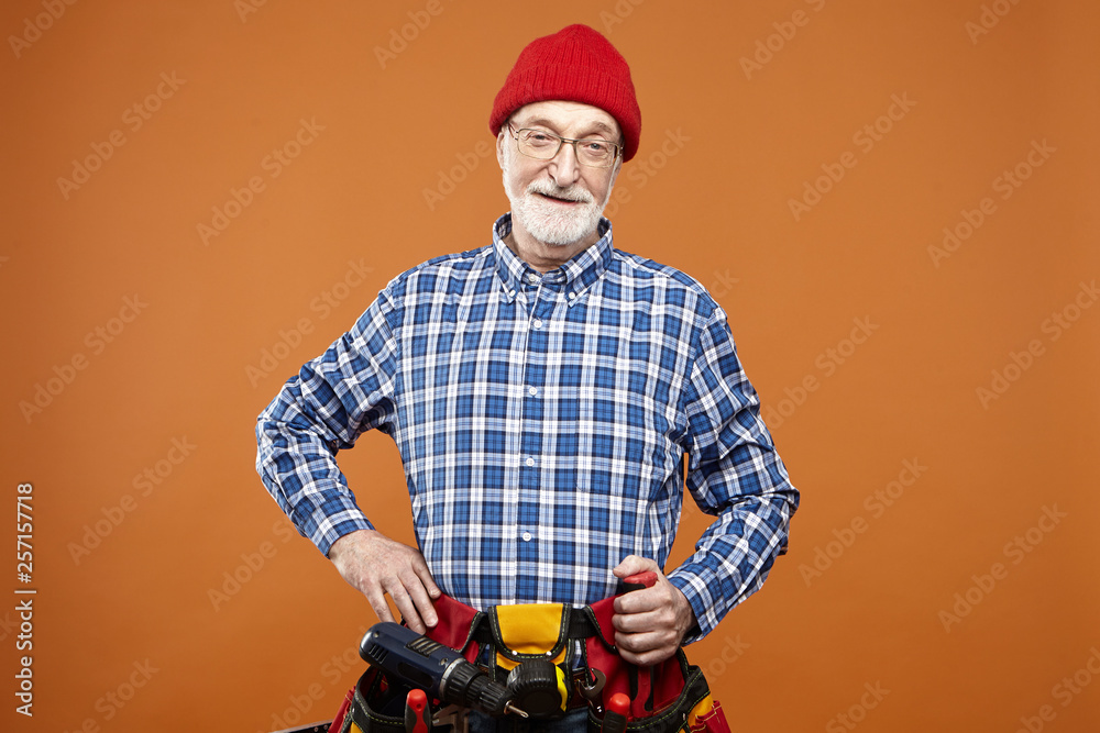 People, aging and occupation concept. Horizontal shot of confident elderly builder with gray beard getting ready for new workday, wearing plaid shirt, red hat and tool belt with all instruments
