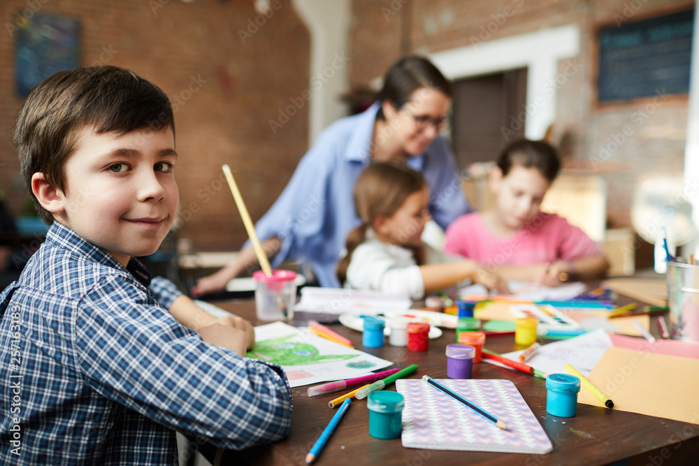 Portrait of smart little boy looking at camera while painting pictures in art class with group of children, copy space Portrait of smart little boy looking at camera while painting pictures in art class with group of children, copy space