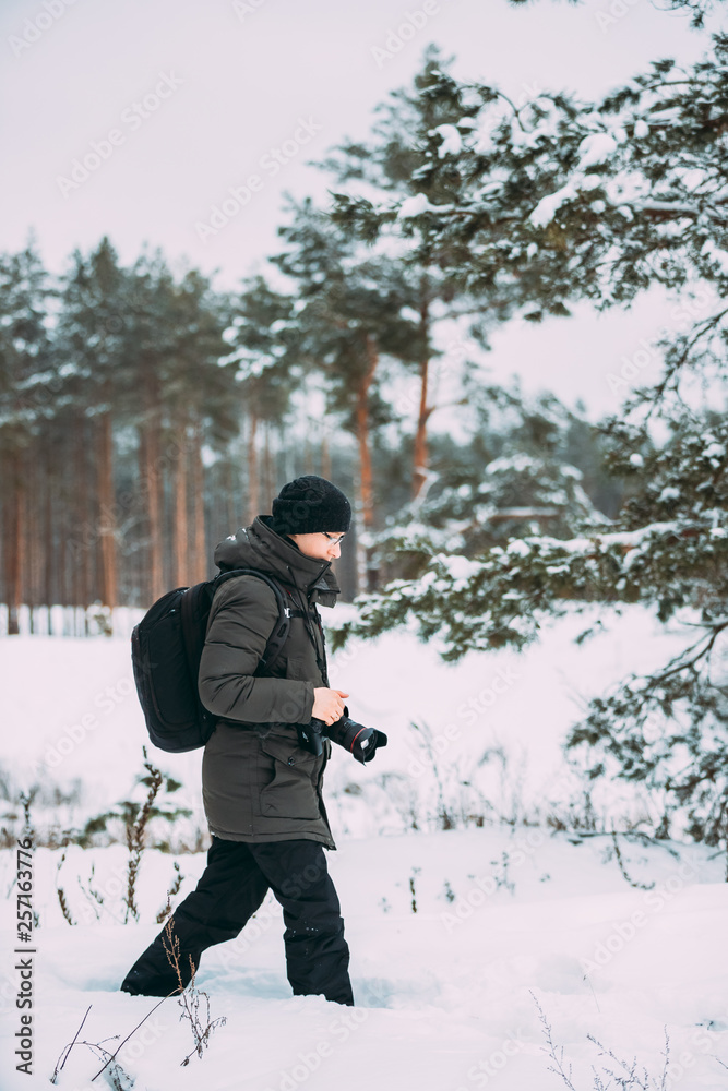 Young Man Backpacker With Photo Camera Taking Photo In Winter Snowy Forest. Active Hobby. Hiker Walking In Snowy Pinewood Forest