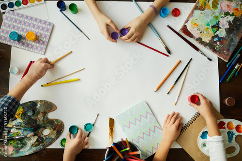 Top view of childrens hands over background with art supplies, paints and brushes over white paper, copy space