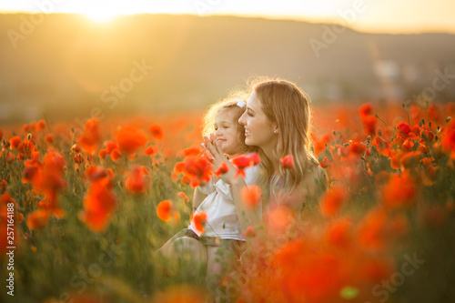 Beautiful smiling baby girl with mother are having fun in field of red poppy flowers over sunset lights, spring time
