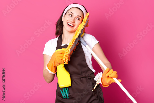 Close up portrait of housewife with good mood, wants to start cleaning her house, has pleasent facial expressions, holds yellow mop and detergent spray oisolated over rose background. Copy space. photo