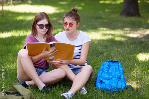 Young girls sitting on grass in park and prepares for classes, wears casual clothes and sunglasses, sits with crossed legs, holds yellow paper folder, read information. Students and education concept.