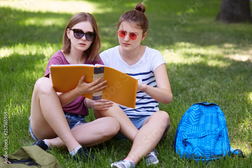 Two young woman study togheter in park, wears casual clothes and sunglasses, read abstracts while prerarig for seminar in unuversity,have concentrated facial eexpressions. Students and education photo