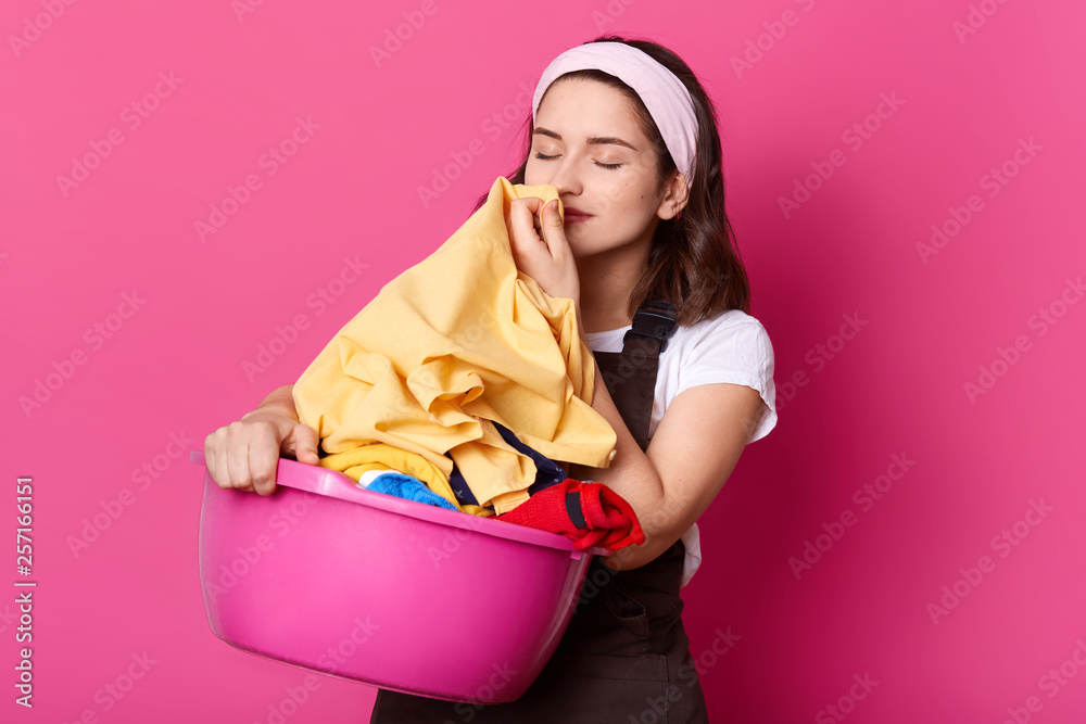 Photo of young attractive woman working at home, wears t shirt, brown apron and hair band, standing with pink basin with clean linen isolated on rose background in photo studio, smells fresh clothes. Photo of young attractive woman working at home, wears t shirt, brown apron and hair band, standing with pink basin with clean linen isolated on rose background in photo studio, smells fresh clothes.