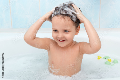 Photographie Little cute girl taking bath, washing her hair with shampoo, looks satisfied beeig in bathtub alone, playing with foam bubbles, looking at camera, Independent infant enjoys being in warm water
