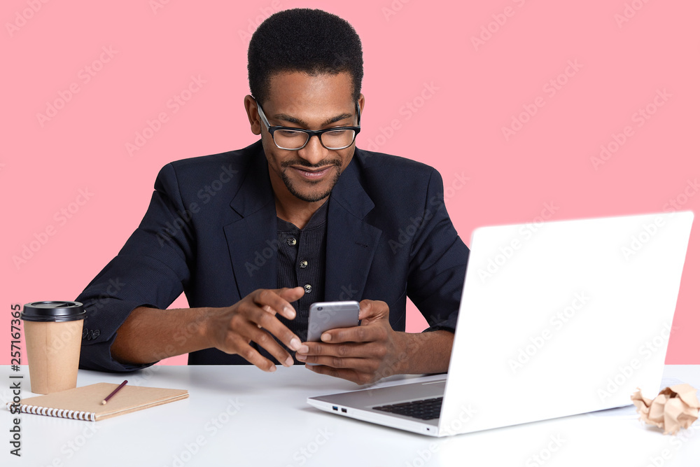 Close up portrait of dark skinned female with modern mobile phone in hands. Handsome black man uses lap top, for working, decides checks social net work while having brake, isolated on pink background Close up portrait of dark skinned female with modern mobile phone in hands. Handsome black man uses lap top, for working, decides checks social net work while having brake, isolated on pink background