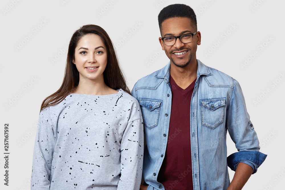 Positive girl and guy of different races poses against white studio wall, smile positively, express their happiness, dressed in casual outfit, enjoy spare time together. People and relation conept. Positive girl and guy of different races poses against white studio wall, smile positively, express their happiness, dressed in casual outfit, enjoy spare time together. People and relation conept.