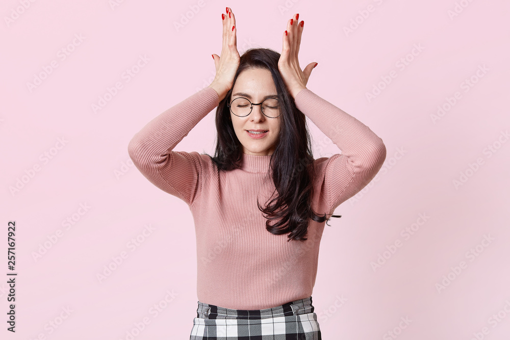 Photo of dissatisfied young dark haired woman, keeps hand on forehead, clenches teeth, dressed in sweater and skirt, adorable female forgets importent thing at home, poses isolated over pink wall.