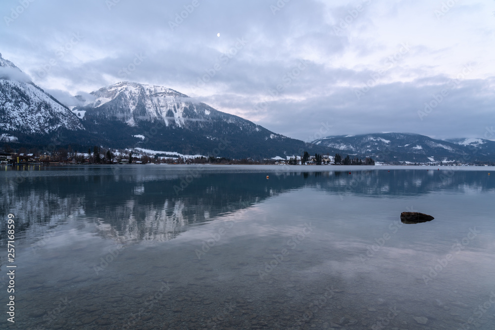 Wolfgangsee mit Spiegelung der schneebedeckten Berge im Hintergrund im klaren Wasser, treibendes Holz, Mond am Himmel
