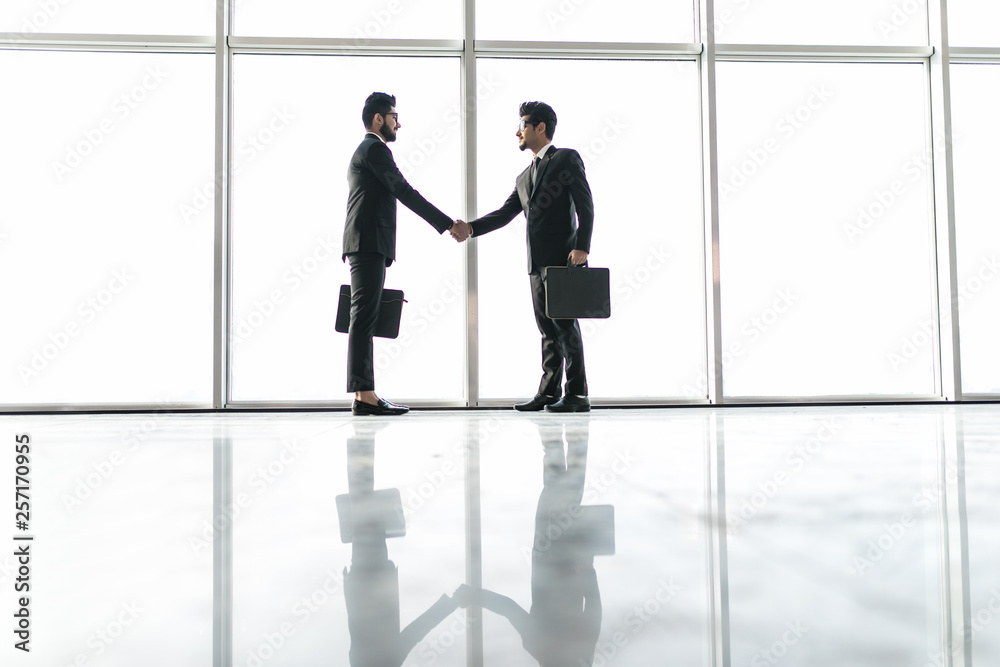 Two young indian businessmen are shaking hands with each other standing against panoramic windows.