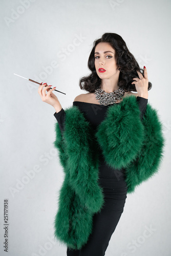 charming graceful pin up girl in a black evening dress and a green fluffy boa in her hands stands and smokes a cigarette with a mouthpiece on a white solid Studio background