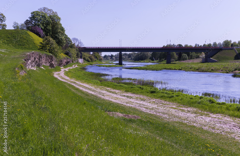 a winding river with a bridge over it; along the edge of the river there is a empty, gravelly rural road; on the left is a hill with a flowering lilac bush a winding river with a bridge over it; along the edge of the river there is a empty, gravelly rural road; on the left is a hill with a flowering lilac bush