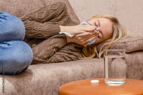 blonde girl sitting on the couch. She has a runny nose, so she blows her nose and wipes away tissue paper. place the medicine and a glass of water on the table next to it. The concept of the disease photo