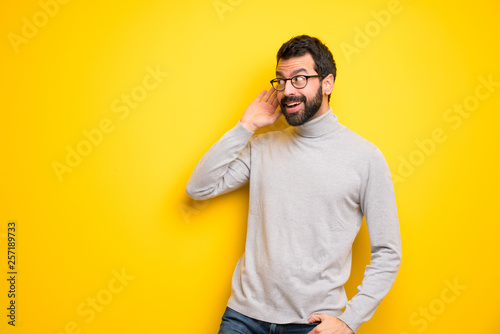 Man with beard and turtleneck listening to something by putting hand on the ear