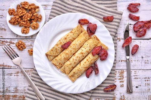 Pancakes, Walnut, Plums. Pancakes with filling on a white plate. Wood background. Rustic style. Rolled up pancakes. Wrapped thin pancakes. Homemade crepes, dried fruits, nuts.
