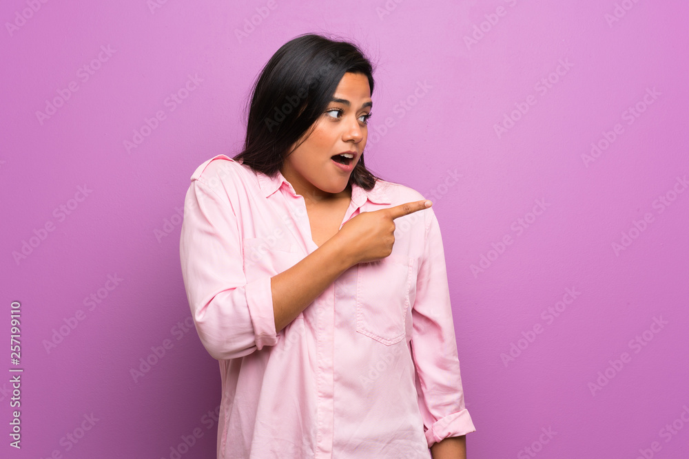 Young Colombian girl over purple wall surprised and pointing side Young Colombian girl over purple wall surprised and pointing side