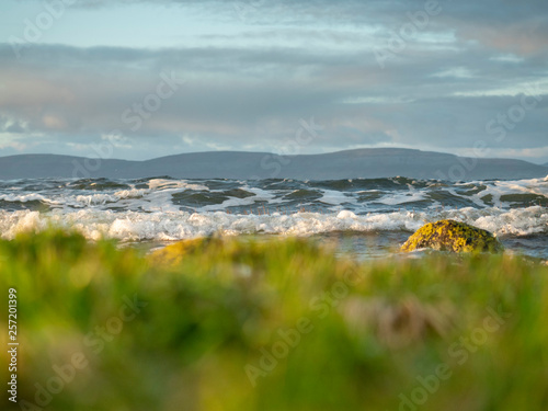 Ocean wave in focus area foreground grass is out of focus, mountains and bluish sky with clouds, Galway bay.