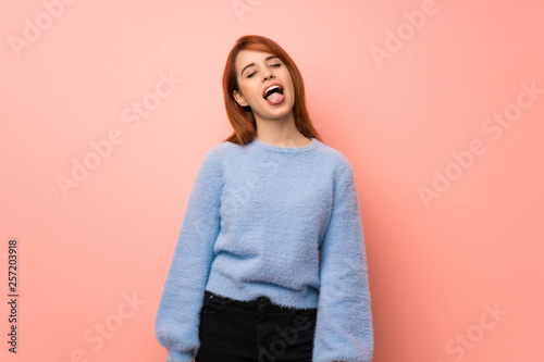 Young redhead woman over pink background showing tongue at the camera having funny look