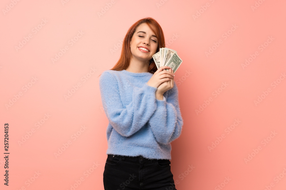 Young redhead woman over pink background taking a lot of money Young redhead woman over pink background taking a lot of money