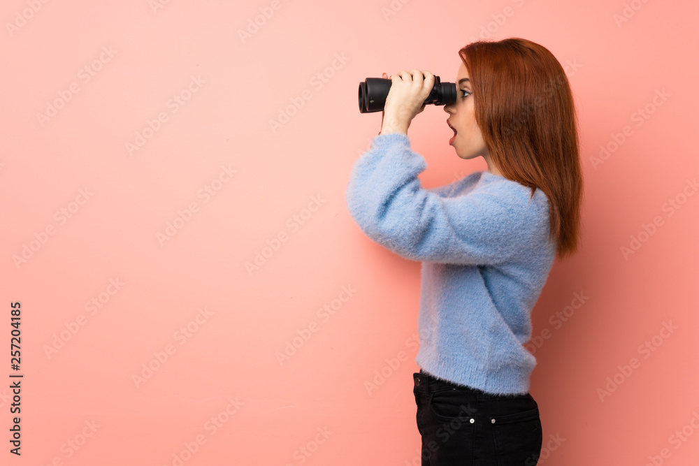Young redhead woman over pink background and looking in the distance with binoculars Young redhead woman over pink background and looking in the distance with binoculars