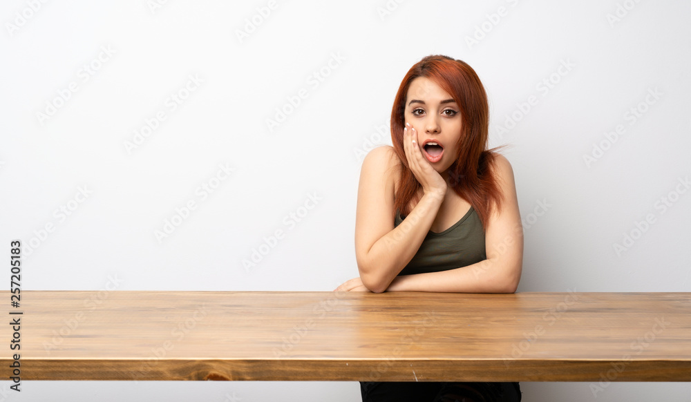 Young redhead woman at desk with surprise and shocked facial expression Young redhead woman at desk with surprise and shocked facial expression